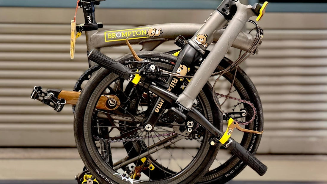 Folded grey Brompton bicycle with custom Wheel Angel carbon wheels and yellow accents indoors.