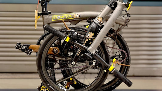 Folded grey Brompton bicycle with custom Wheel Angel carbon wheels and yellow accents indoors.